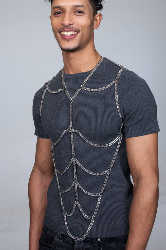 the picture shows a young male model wearing a shiny silver body chain fashion accessory made out fof strong cuban chains. the accessorie is fashionably placed over a gray T-shirt. The models handsome face is smiling