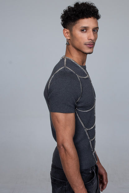 The picture shows a handsome muscular male model from the side wearing a street style outfit in black and gray. On top of the T-shirt the model wears a fashionable body chain accessory by the fashion brand Lorand Lajos
